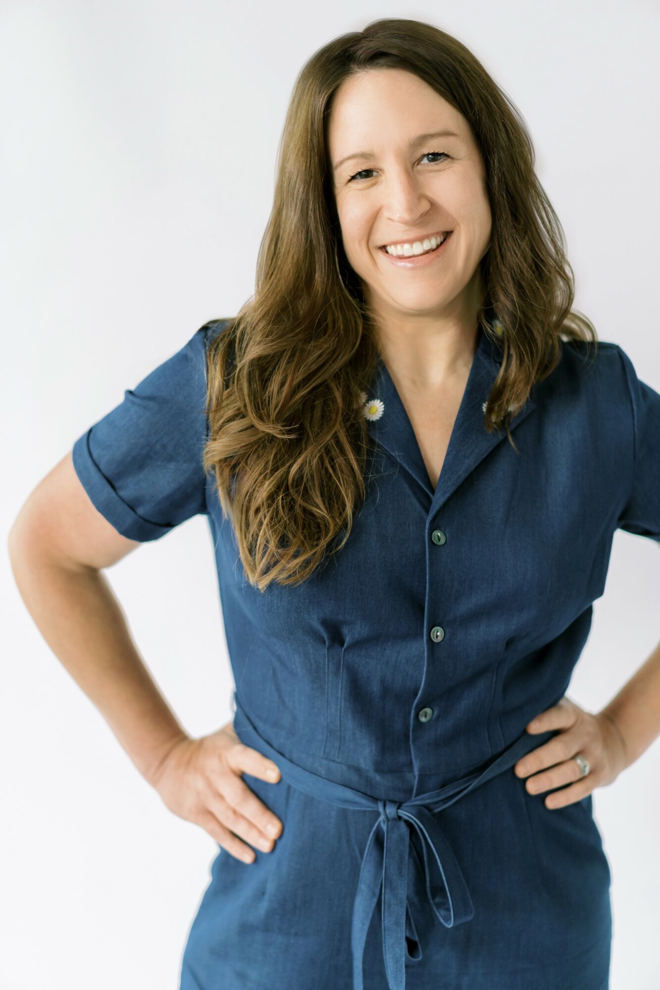 Woman with long wavy hair smiling, wearing a blue dress with a tied waist, hands on hips.