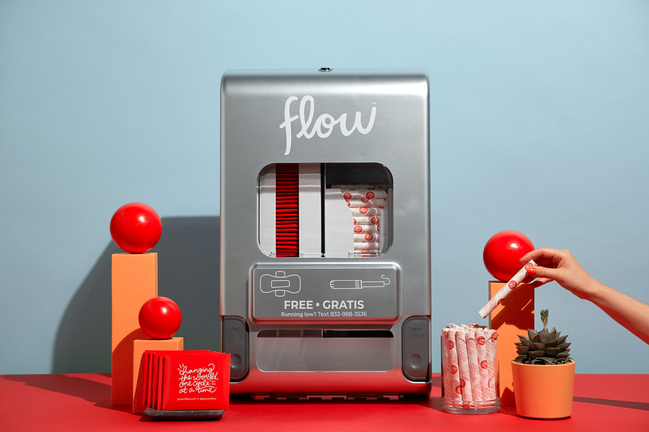 Juice dispenser with red and white striped design, labeled 'flow,' surrounded by red and orange decorative objects and a hand holding a straw.
