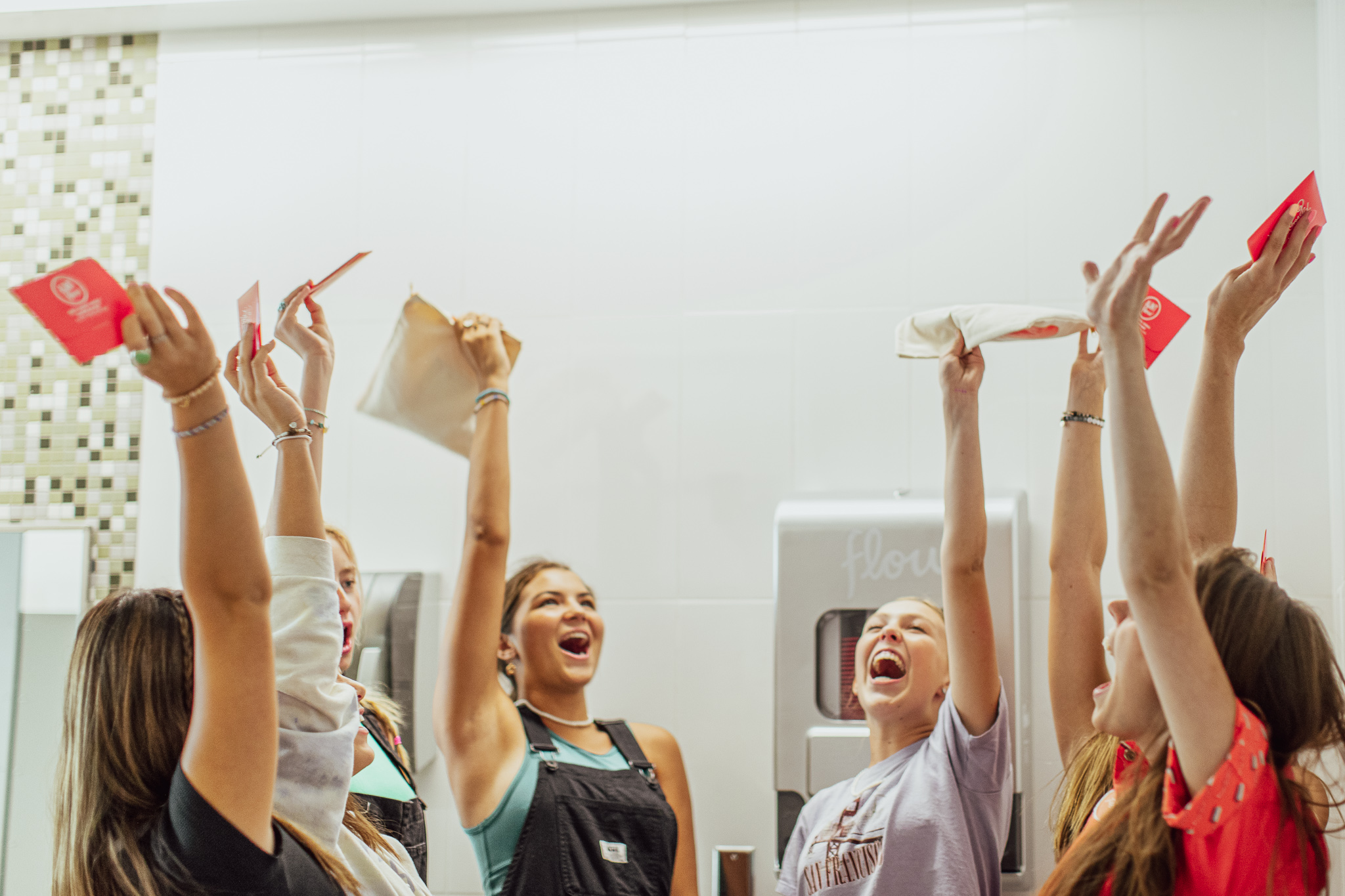 Group of people celebrating with raised hands, holding red envelopes and paper rolls, smiling and joyful.