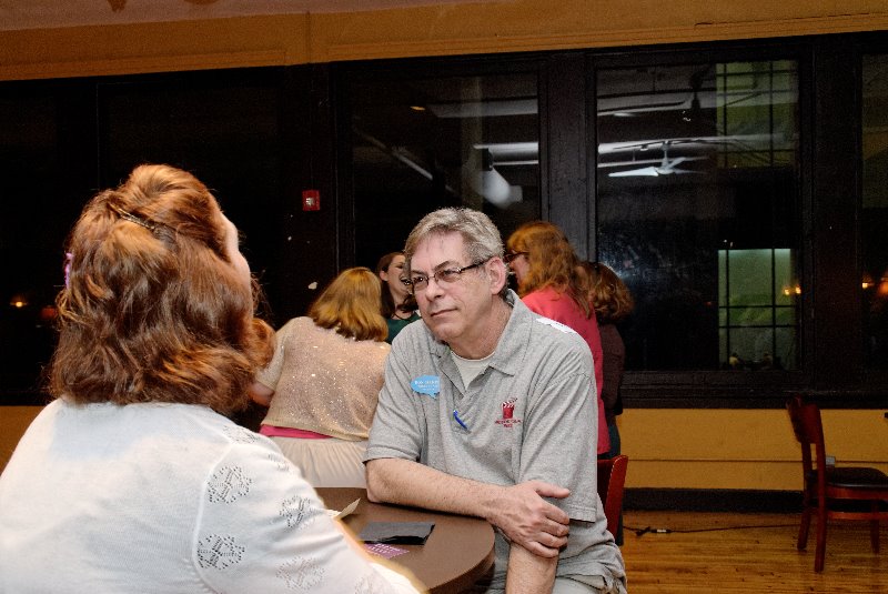 Two people seated at a table engaged in conversation in a room with large windows and other people in the background.