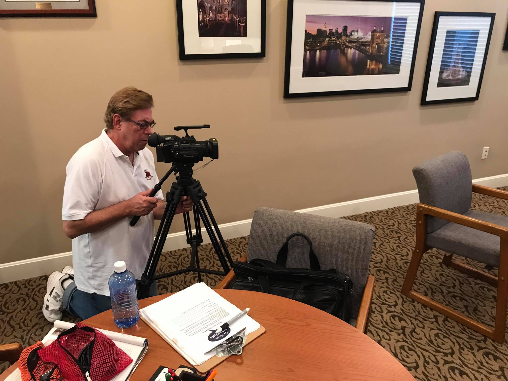 Man operating a camera on tripod in a room with framed pictures on the wall.