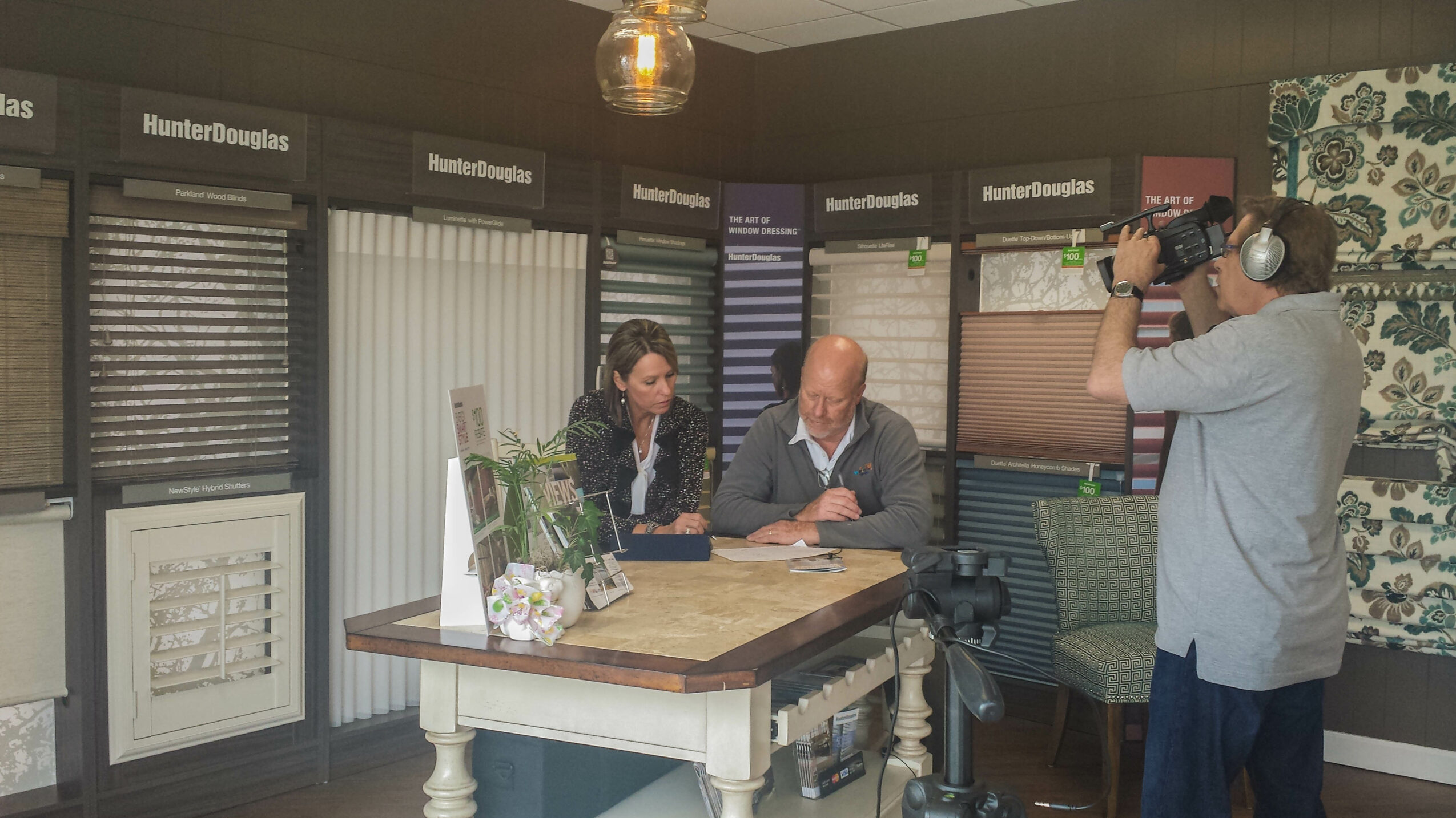 Two people sit at a table with samples of window blinds displayed behind them, while a person films them with a camera.
