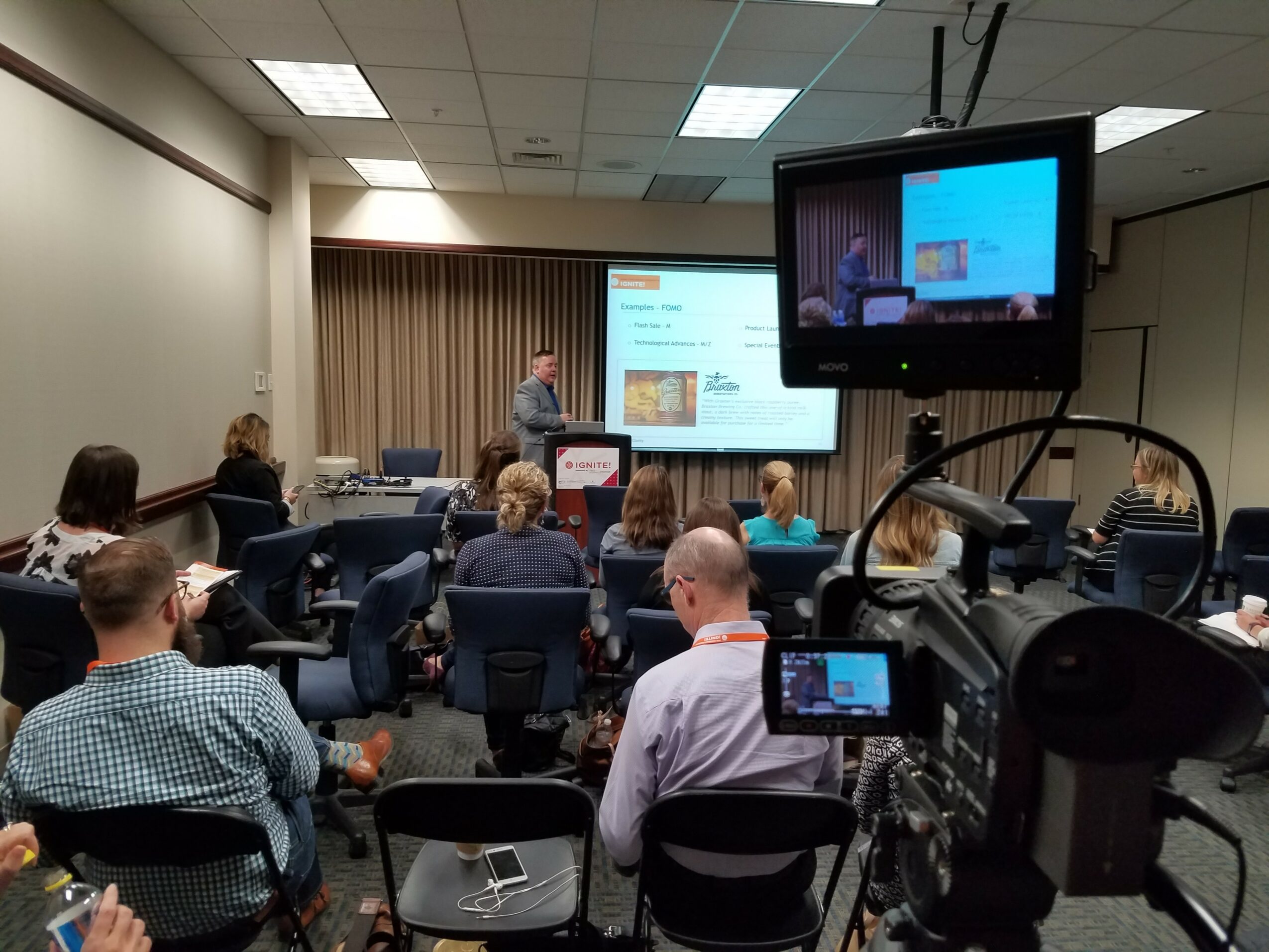 Conference room with a presenter at a podium, audience seated facing screens, one large monitor displaying a presentation slide.