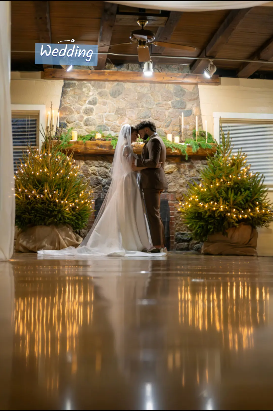 Bride and groom kissing on stage with wedding sign and decorated plants, indoor venue with wooden ceiling.
