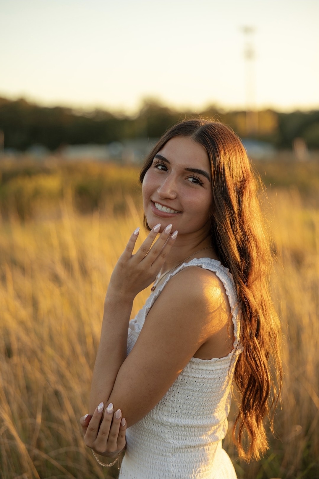 Young woman with long hair smiling outdoors in a field during sunset, wearing a sleeveless top.