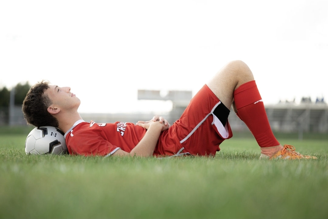 Child lying on grass with soccer ball, wearing red sports uniform, smiling with eyes closed.