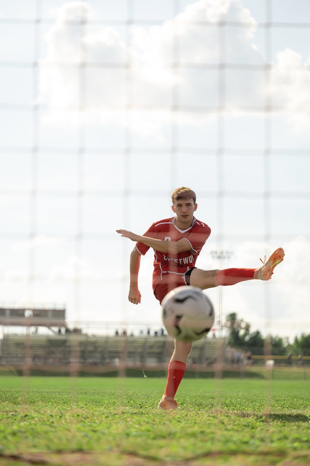 Soccer player in red uniform kicking a ball on a grassy field with a cloudy sky background.