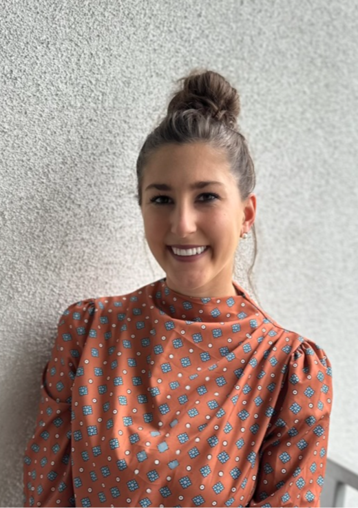 Young woman with a bun hairstyle smiling, wearing a patterned blouse, standing against a textured wall.