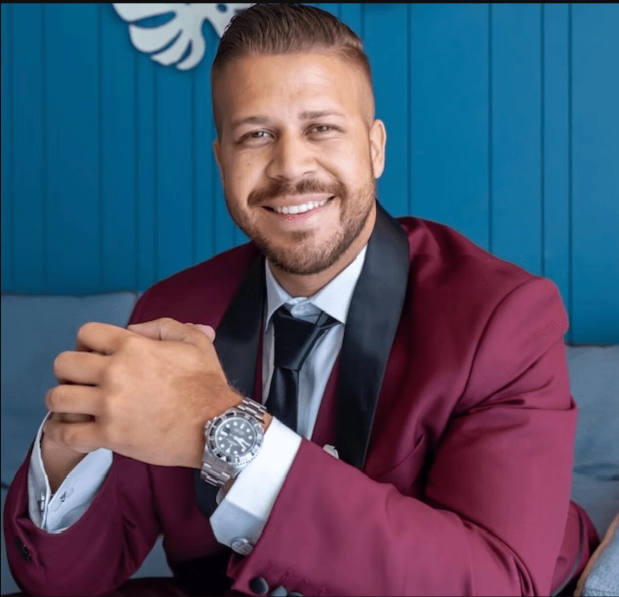 Man with a beard in a maroon suit, smiling, sitting with hands clasped, blue background, wearing a watch.