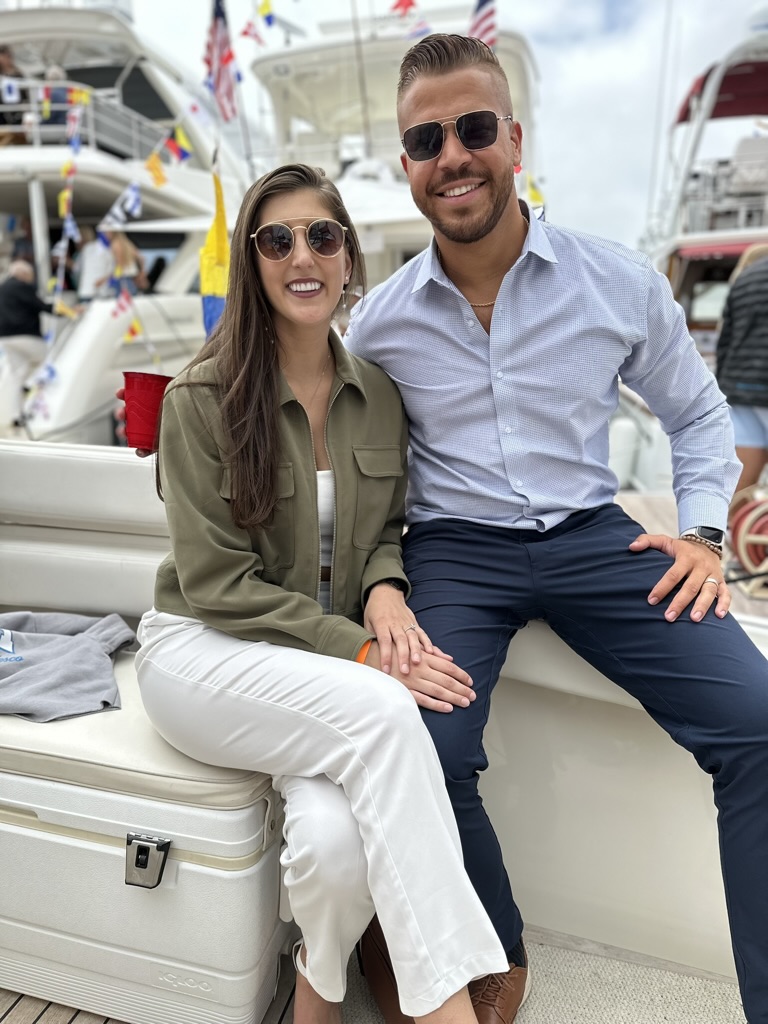 A smiling man and woman sit on a boat dock, with yachts and flags in the background.