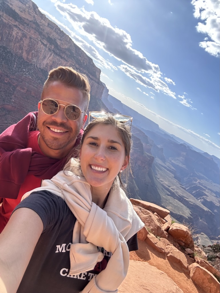 Two people smiling at the camera with a canyon and sky in the background.