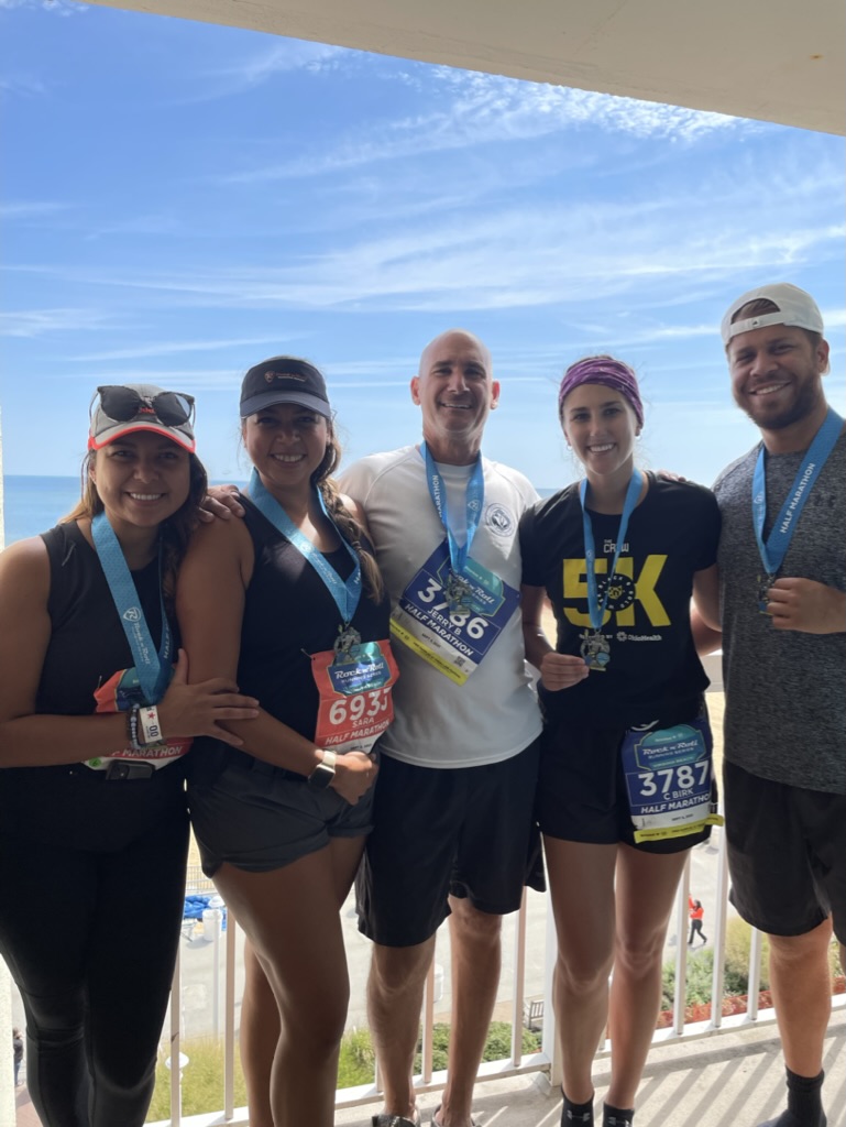 Five people standing outdoors on a balcony with a beach and blue sky in the background, smiling and wearing athletic clothing and medals.