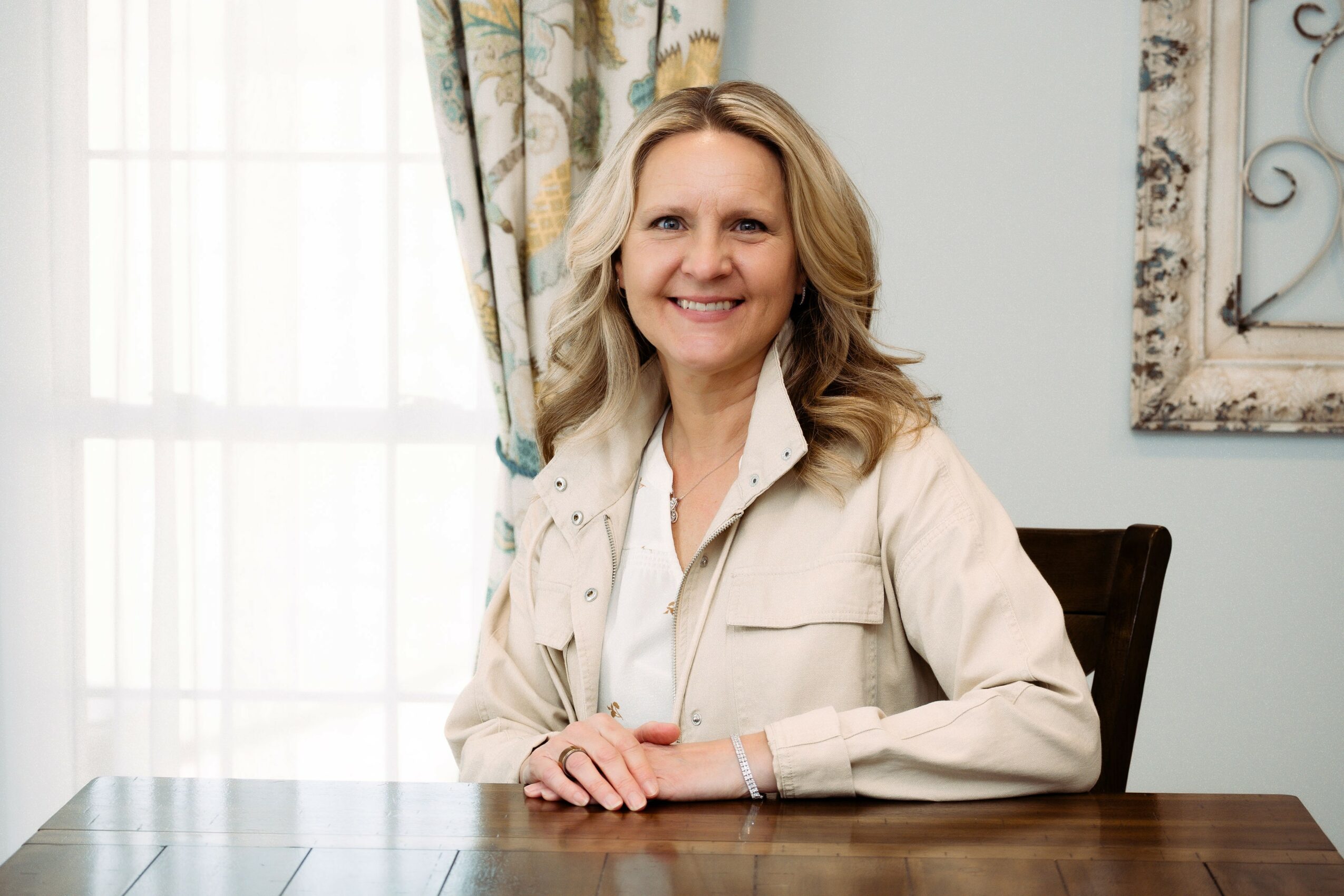 Smiling woman with blonde hair sitting at a wooden table in a bright room with curtains and a decorative wall piece.