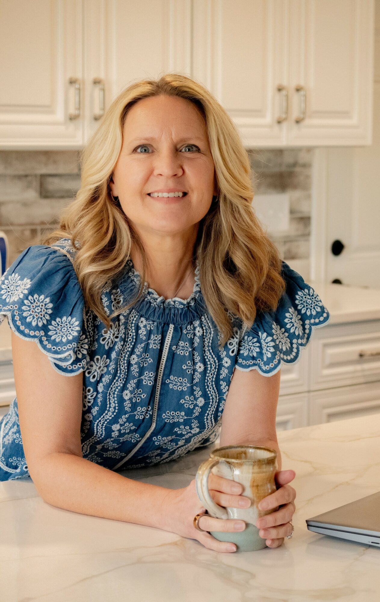 Woman with blonde hair smiling in a kitchen, holding a glass jar, wearing a blue patterned blouse.