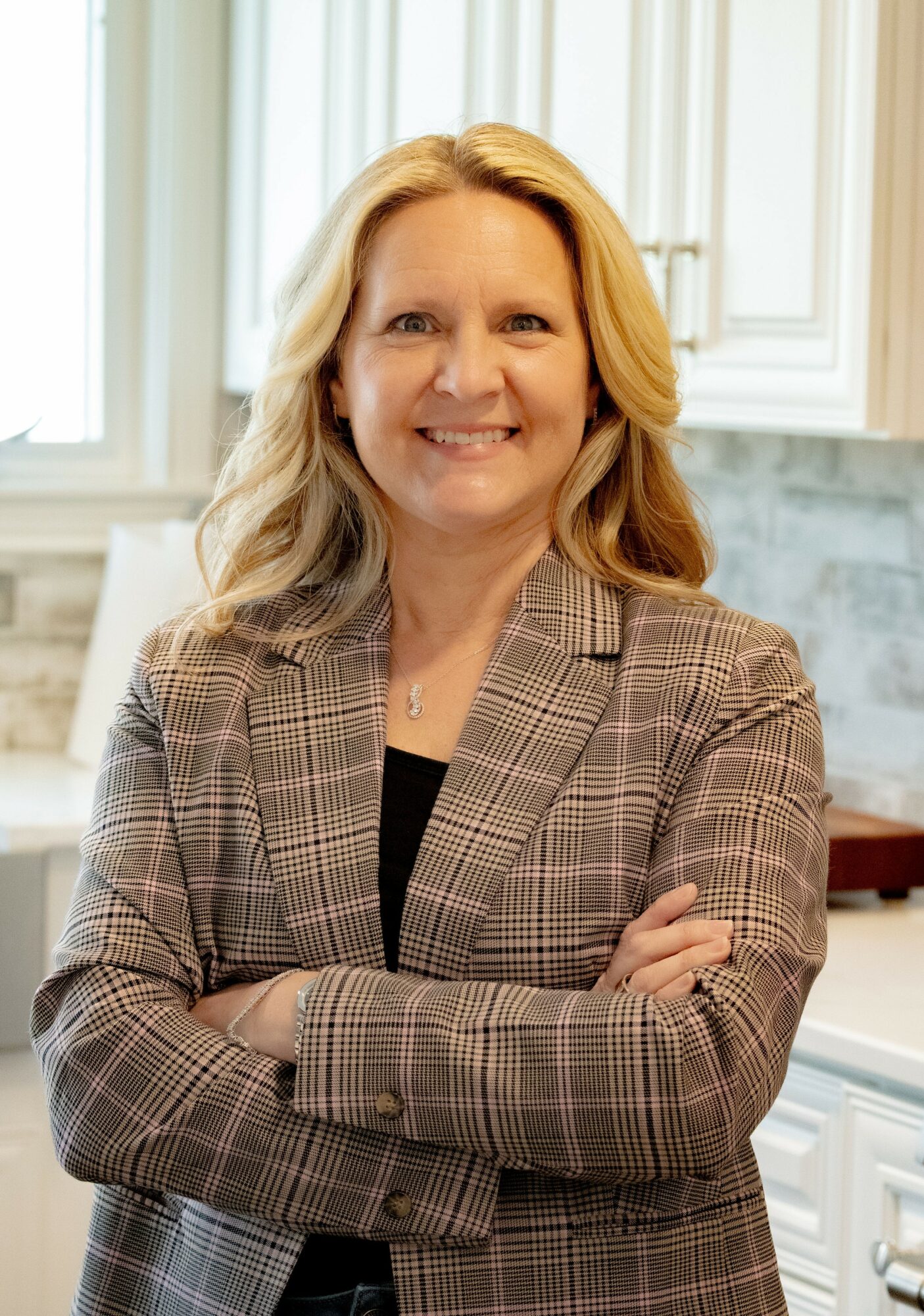 Woman with blonde hair smiling, arms crossed, in a kitchen with white cabinets and marble backsplash.