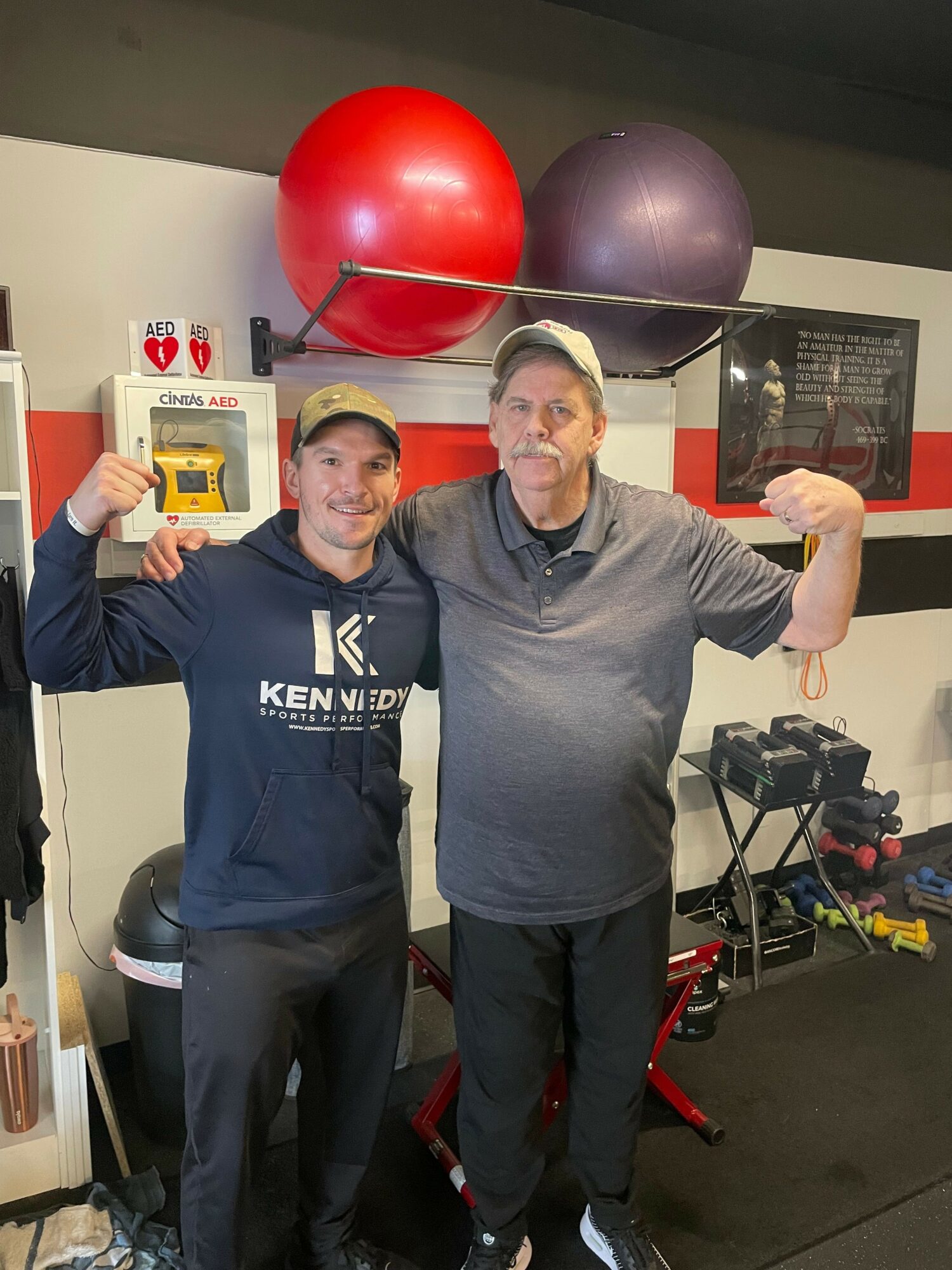 Two men in a gym, flexing their arms, standing in front of exercise balls and equipment.