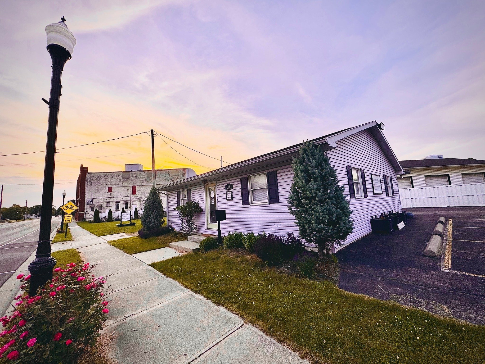 Street view with a house, trees, sidewalk, streetlamp, and sky in the background.