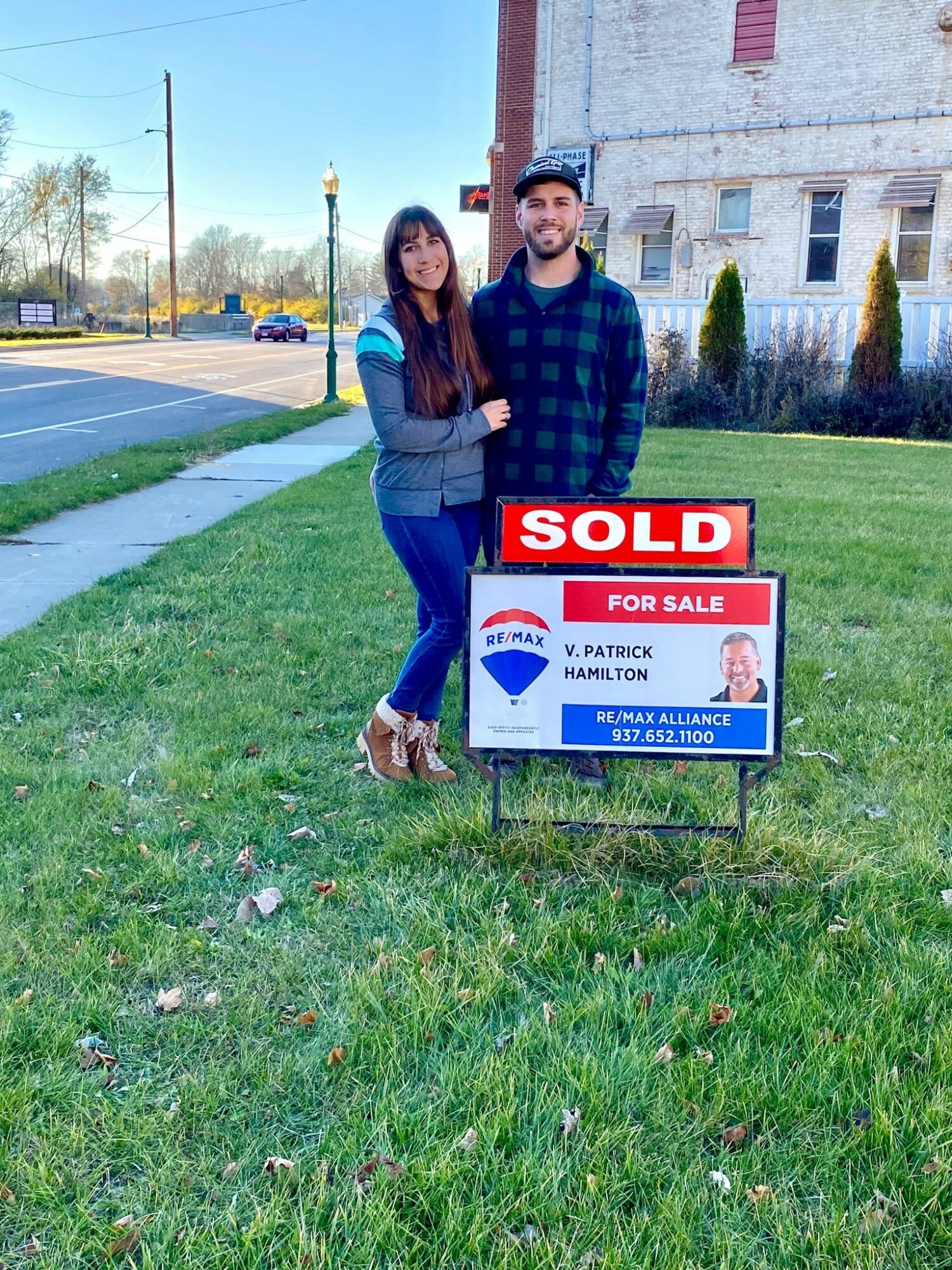 Two people standing on grass next to a sold sign in front of a building, with a street and trees in background.