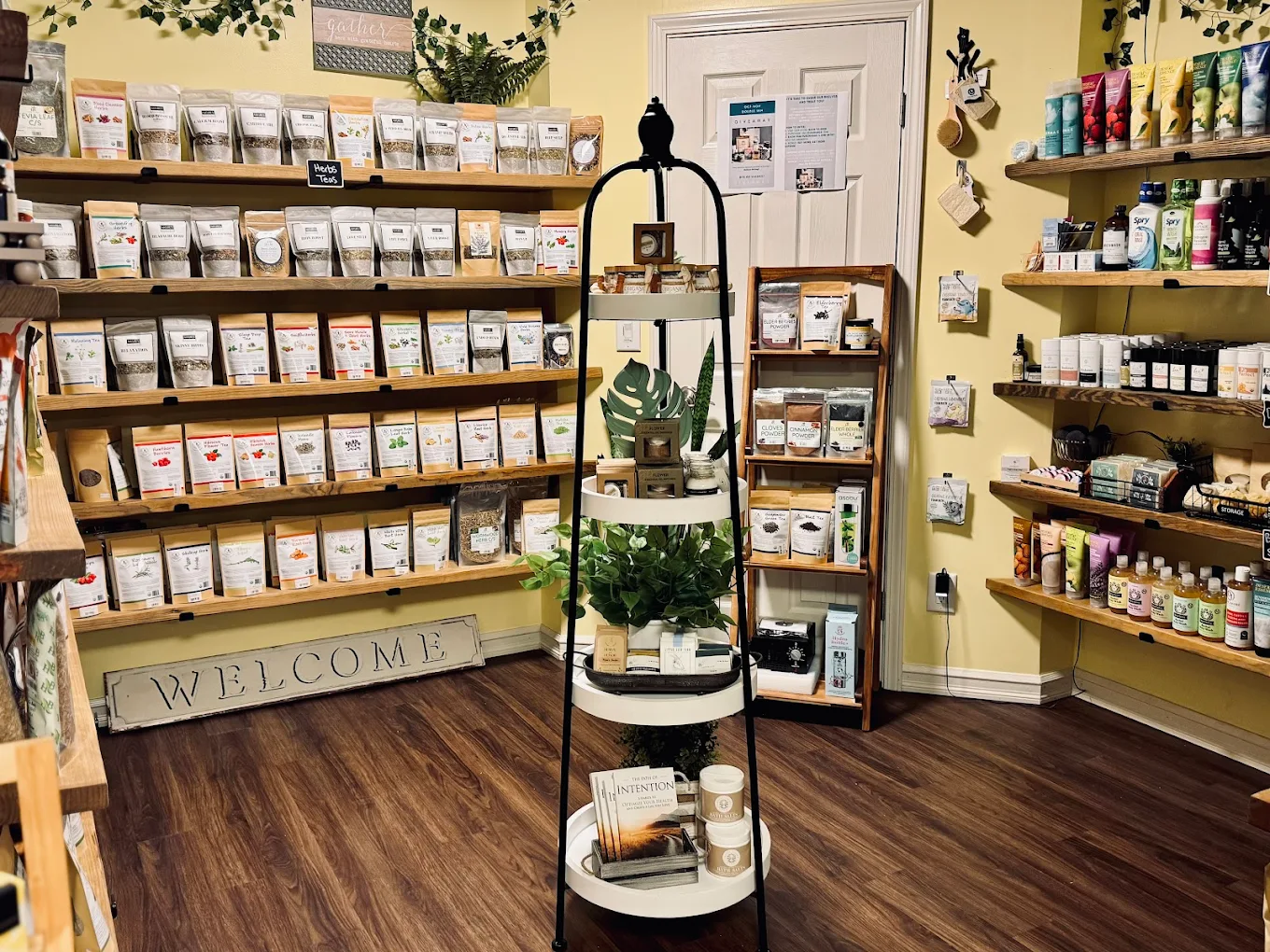 Interior of a store with shelves of products, a welcome sign, and a black metal plant stand with potted plants.