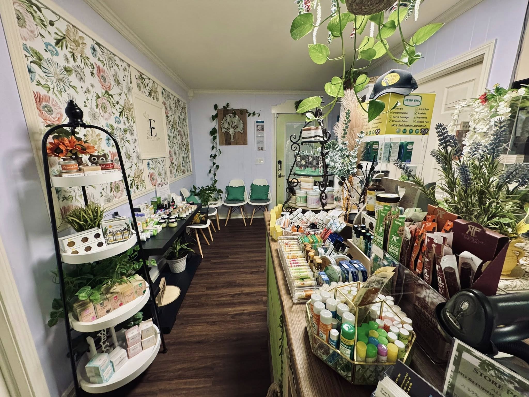 Interior of a store with shelves, plants, and a floral wall, displaying various products and seating area at the back.