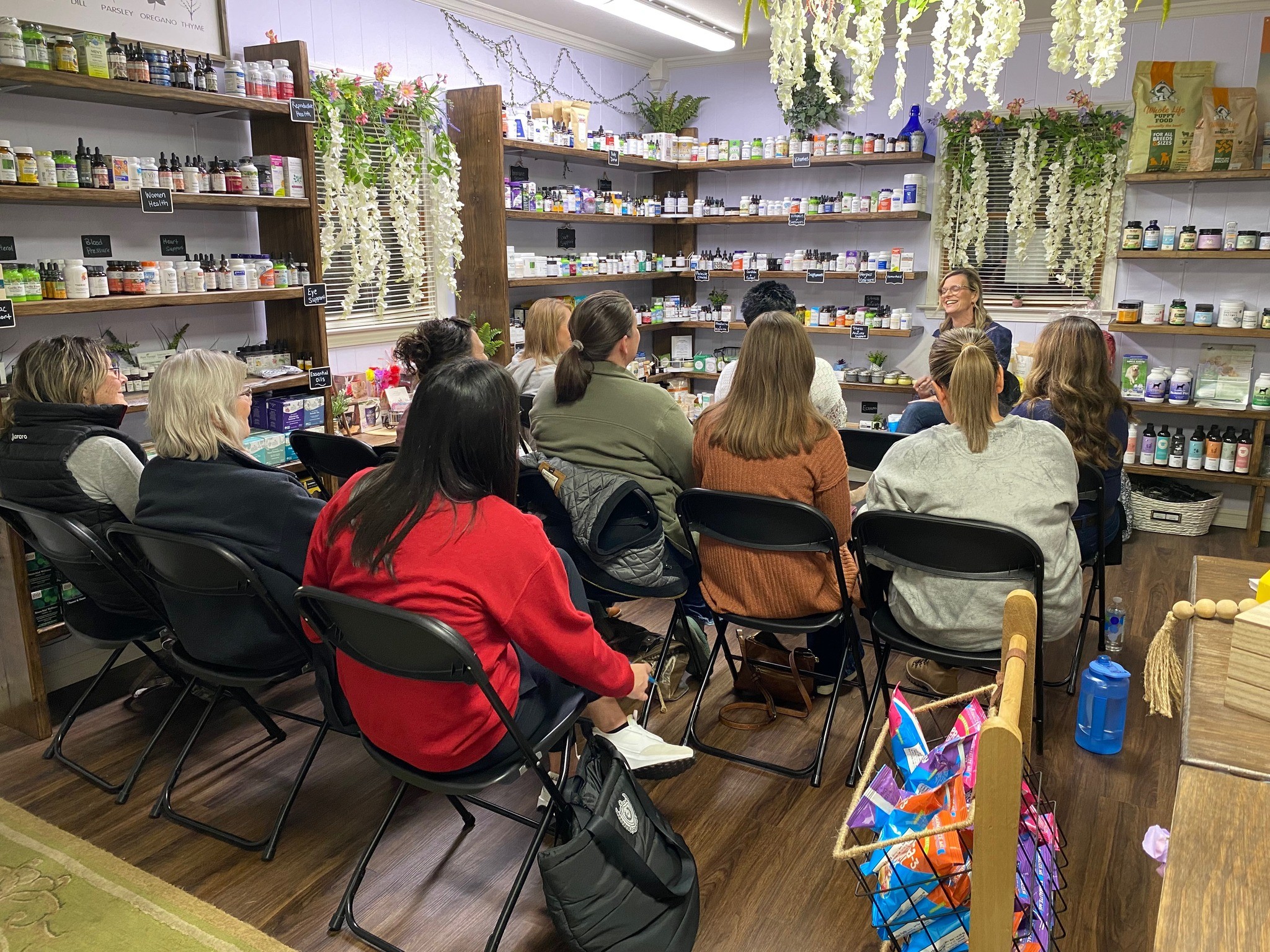 Group of people seated in a room with shelves and hanging plants, listening to a speaker at the front.