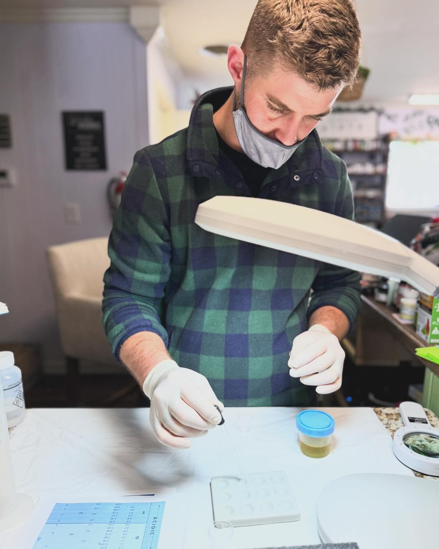 Young man wearing a mask and gloves, looking at a container on a table in a laboratory or workspace.