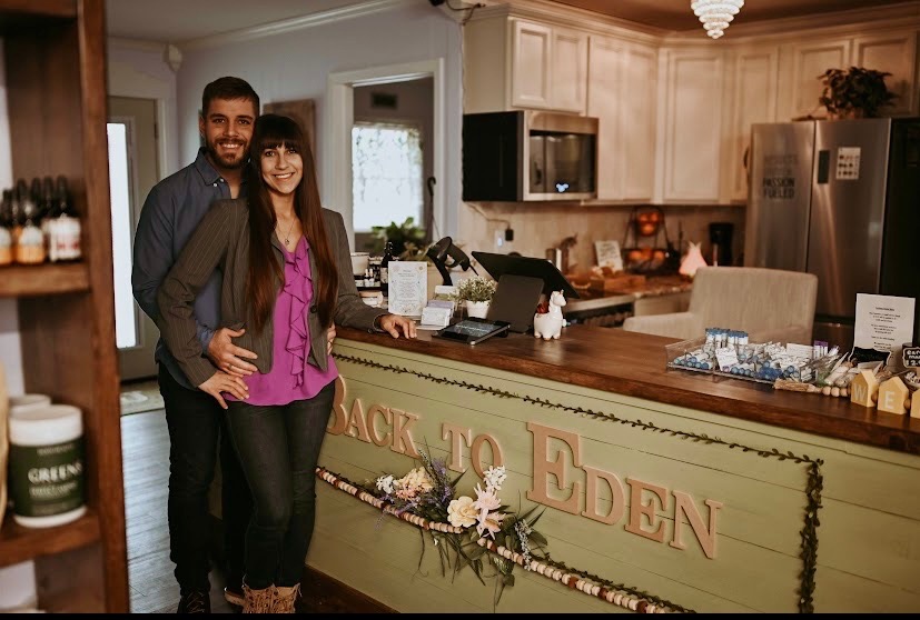 Two people stand behind a decorated counter in a cozy kitchen, smiling at the camera.