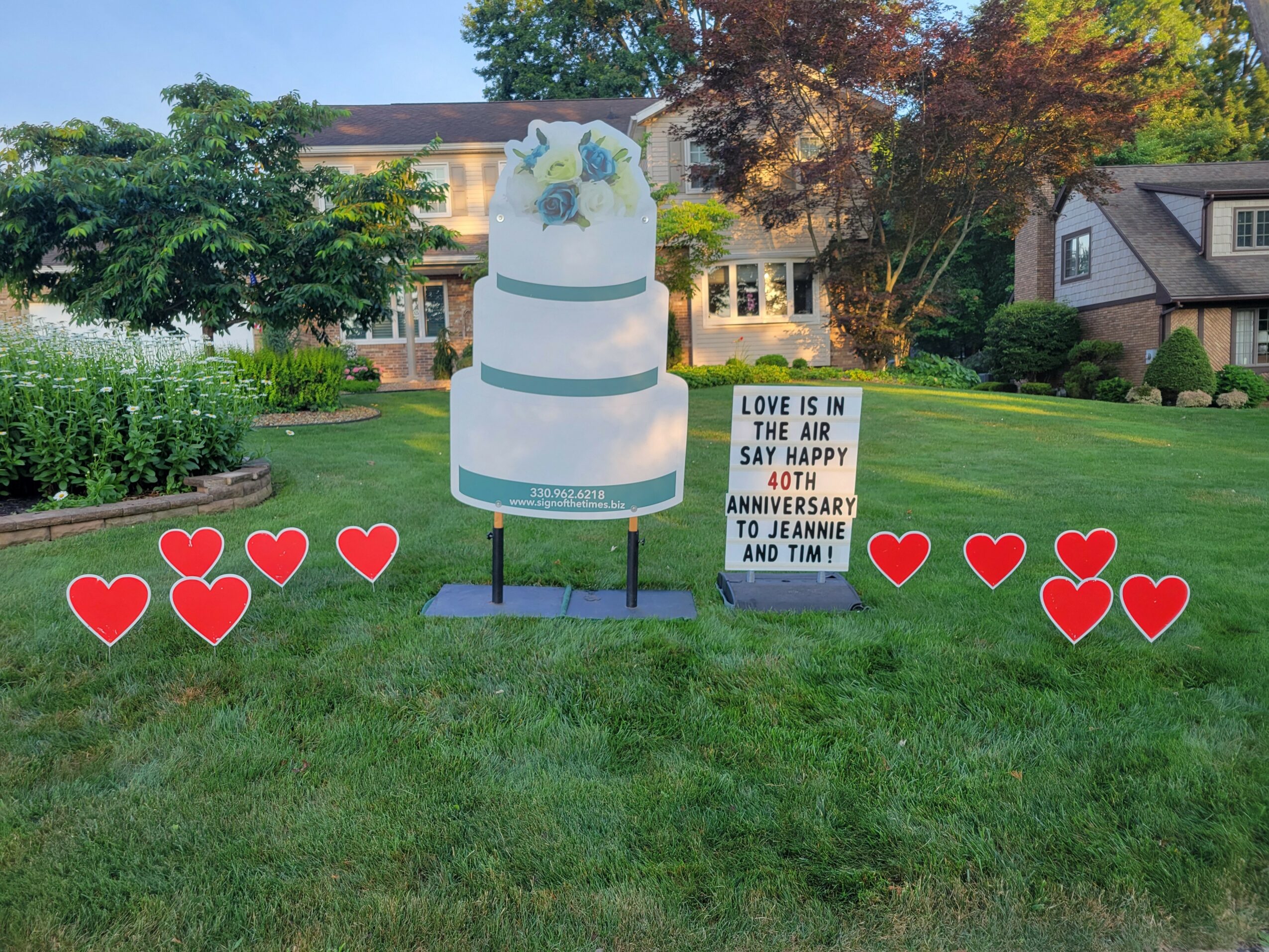 Outdoor wedding display with a cake cutout, a sign, and red hearts on grass with trees and houses in background.