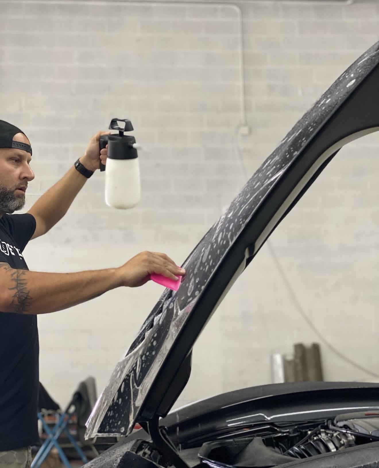 Man cleaning car windshield with spray bottle and cloth in garage, with car hood open.