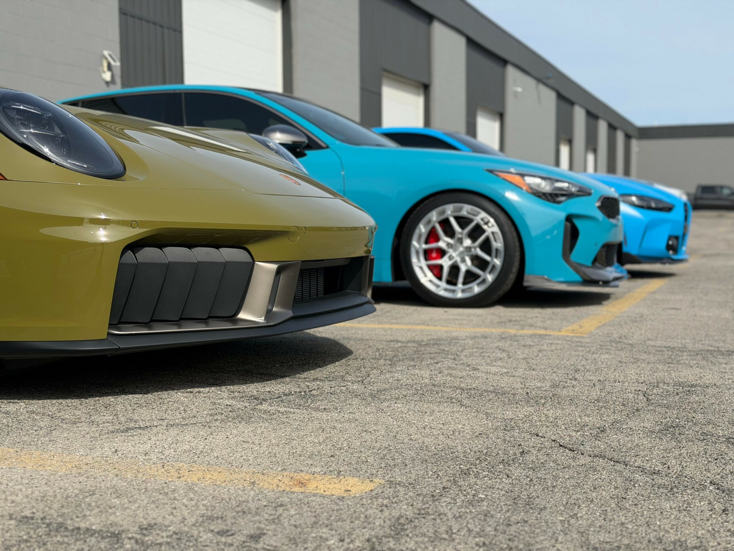 Three sports cars parked in a lot outside a building, with the front car in yellow, the middle in blue, and the last in blue.