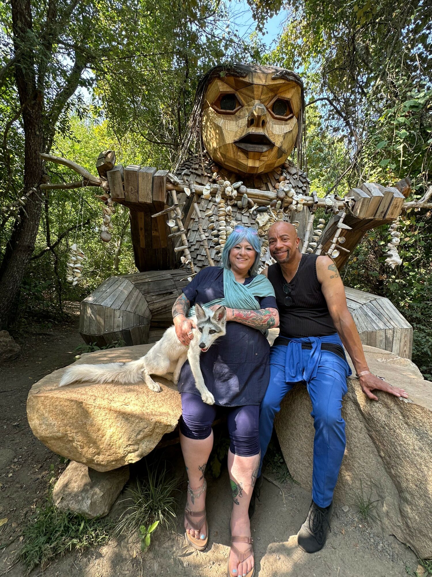 Two people and a dog sitting on rocks in front of a large wooden sculpture of a face with glasses, surrounded by trees.
