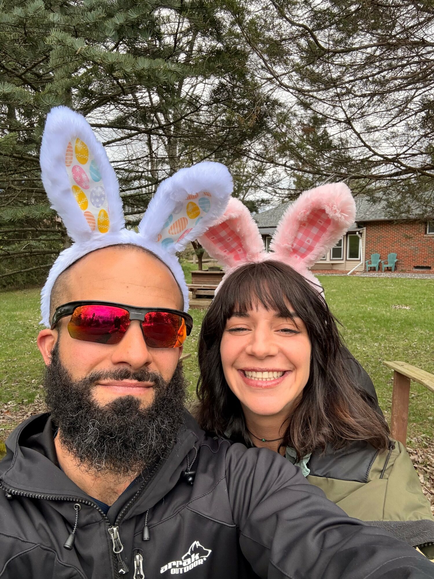 Two people wearing bunny ears headbands smiling outdoors with trees and a building in the background.