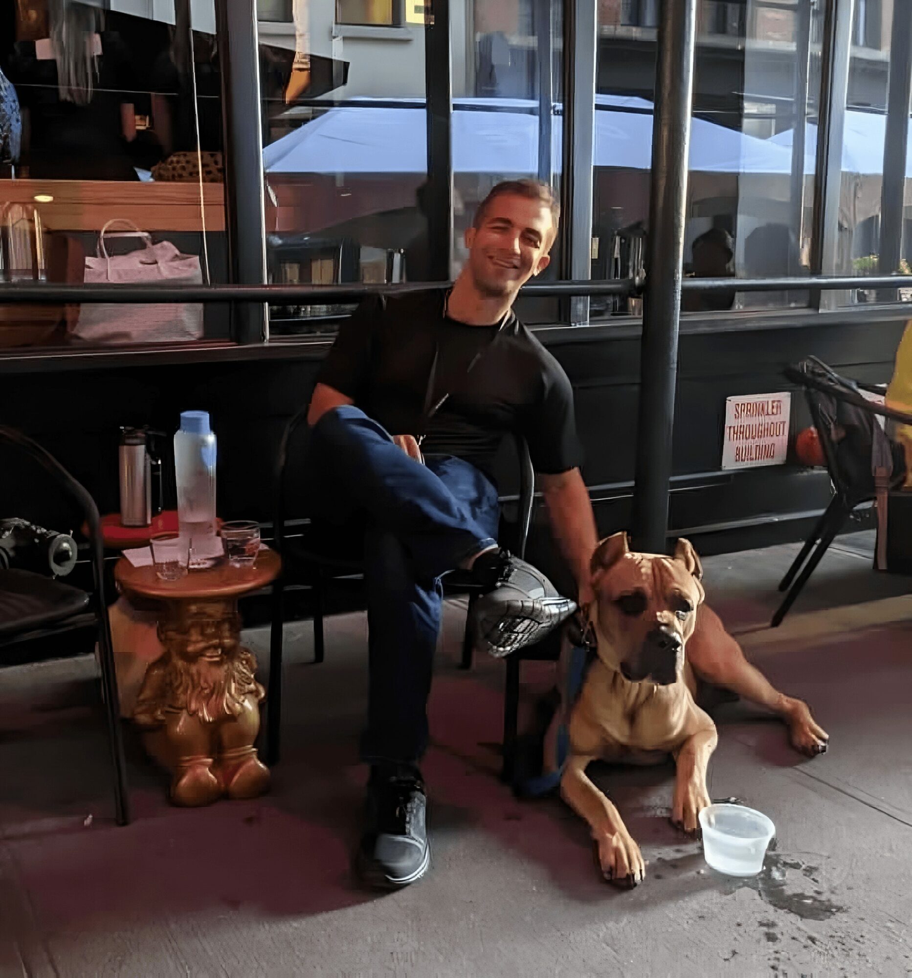 Man sitting on a chair with a large dog lying beside him outside a building with glass windows.