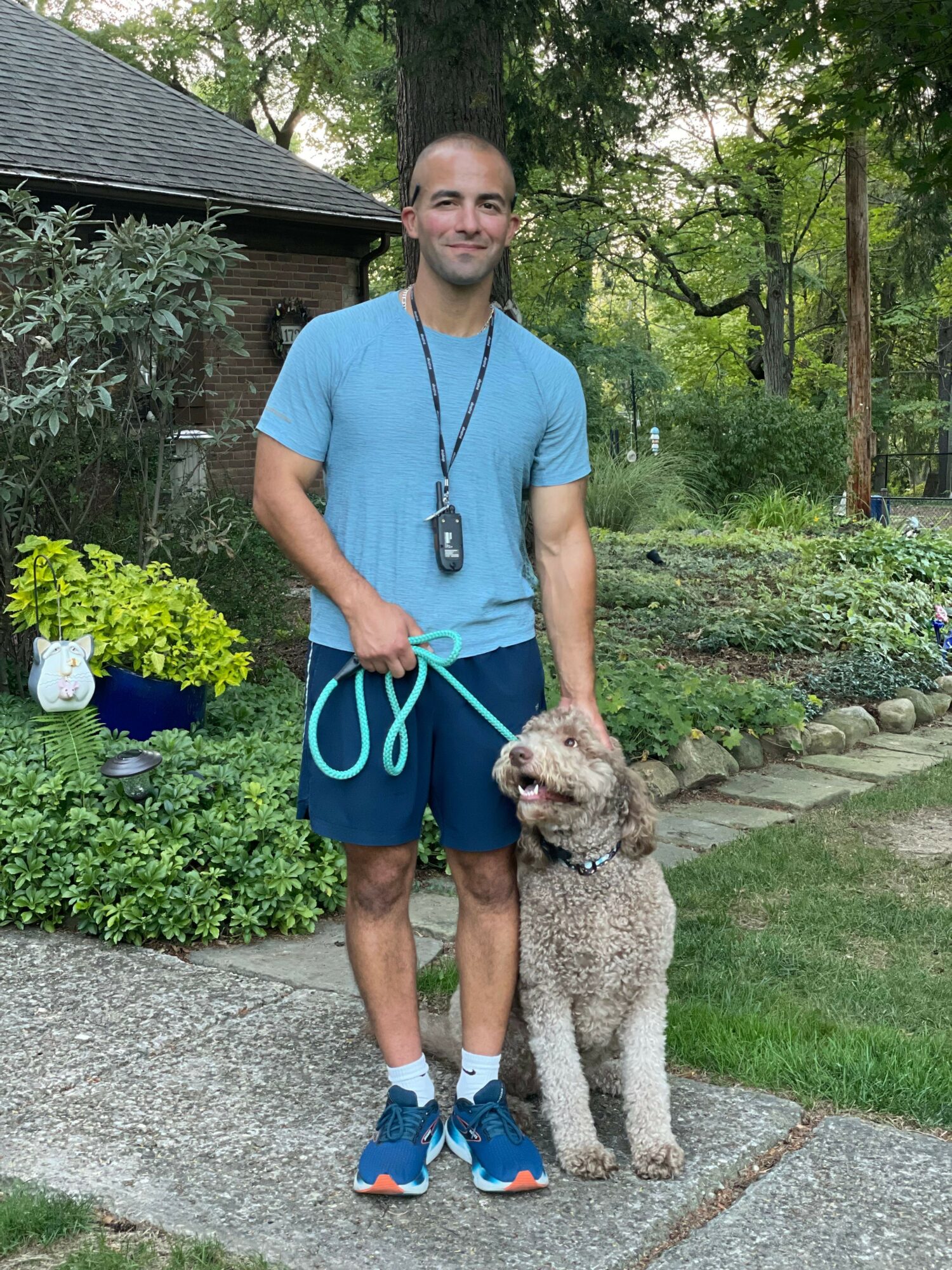 Man in blue shirt and shorts standing outdoors with a dog on a leash, surrounded by greenery and garden plants.