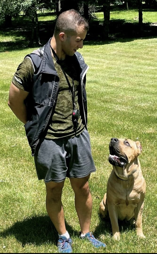 Young man standing outdoors with a dog sitting on grass, trees in background, sunny day.