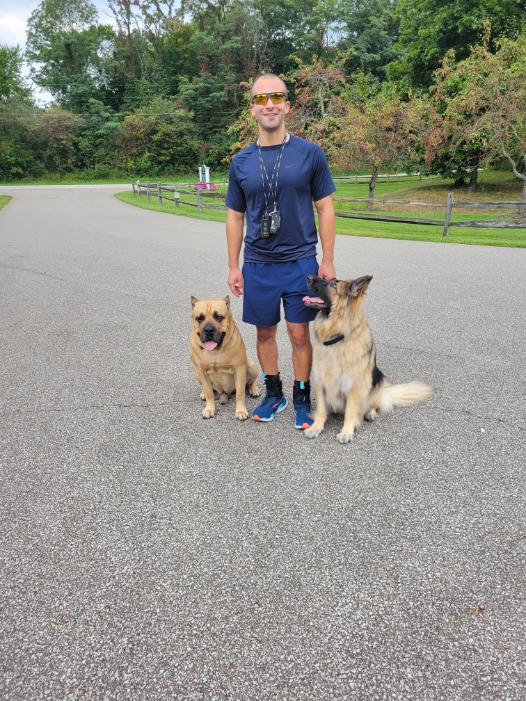 Man standing outdoors with two dogs on a paved path, trees and greenery in background.