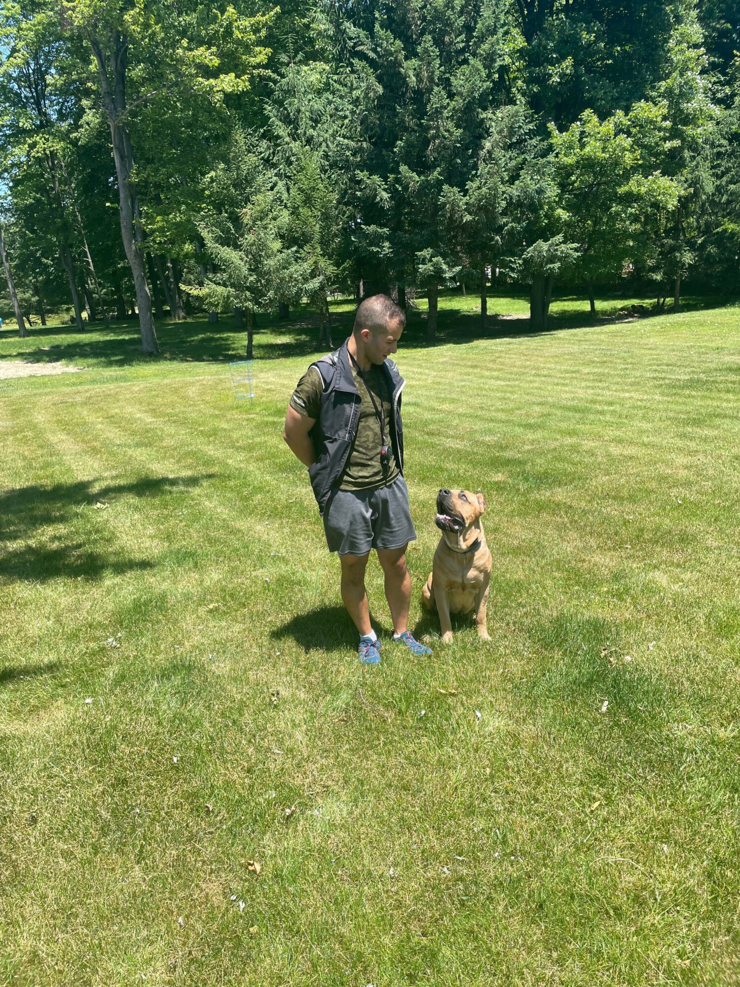 Man standing on grass with a dog sitting beside him, trees in background, sunny day.