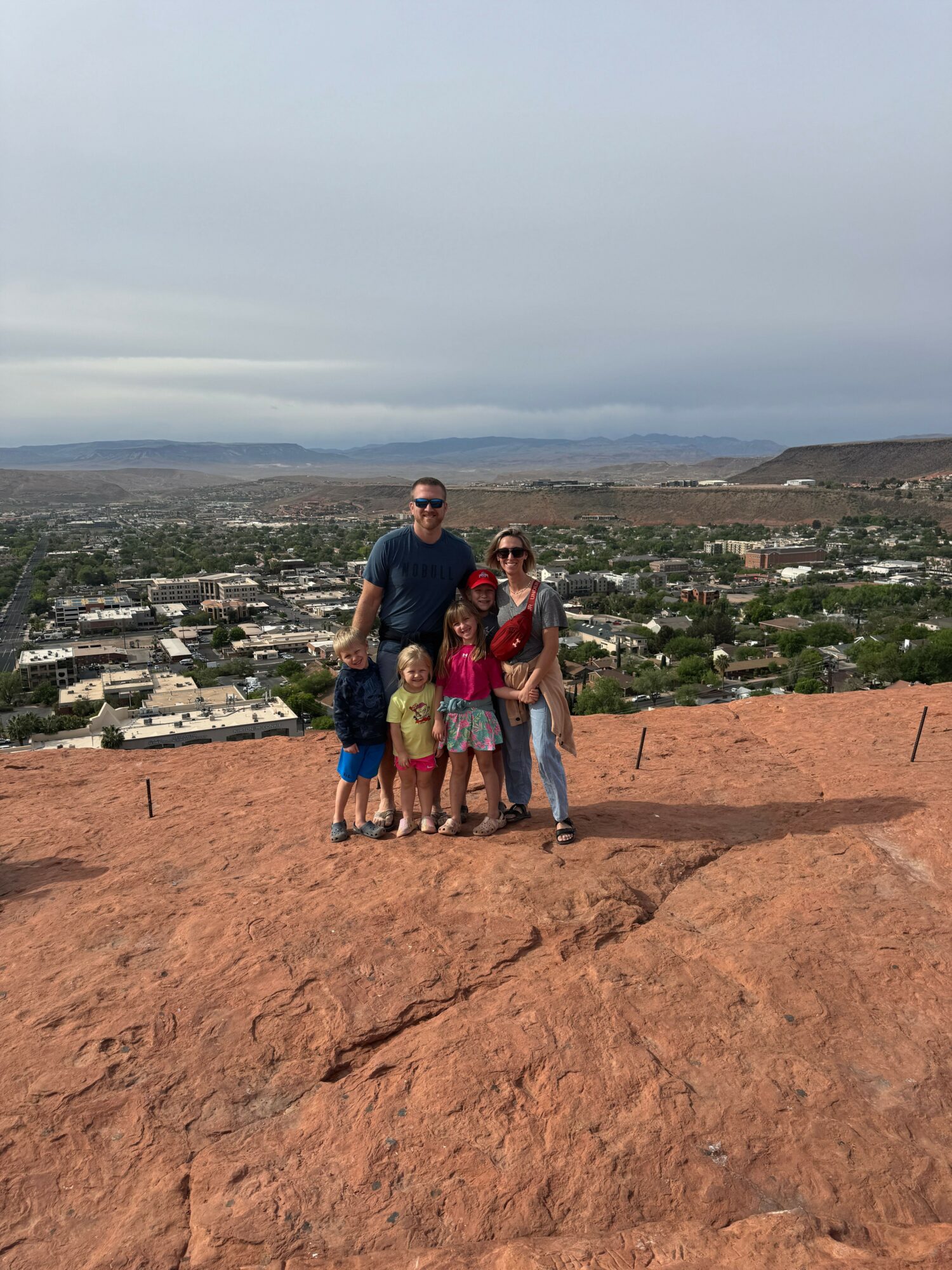 Family of five standing on a red rocky hill with a cityscape and hills in the background.