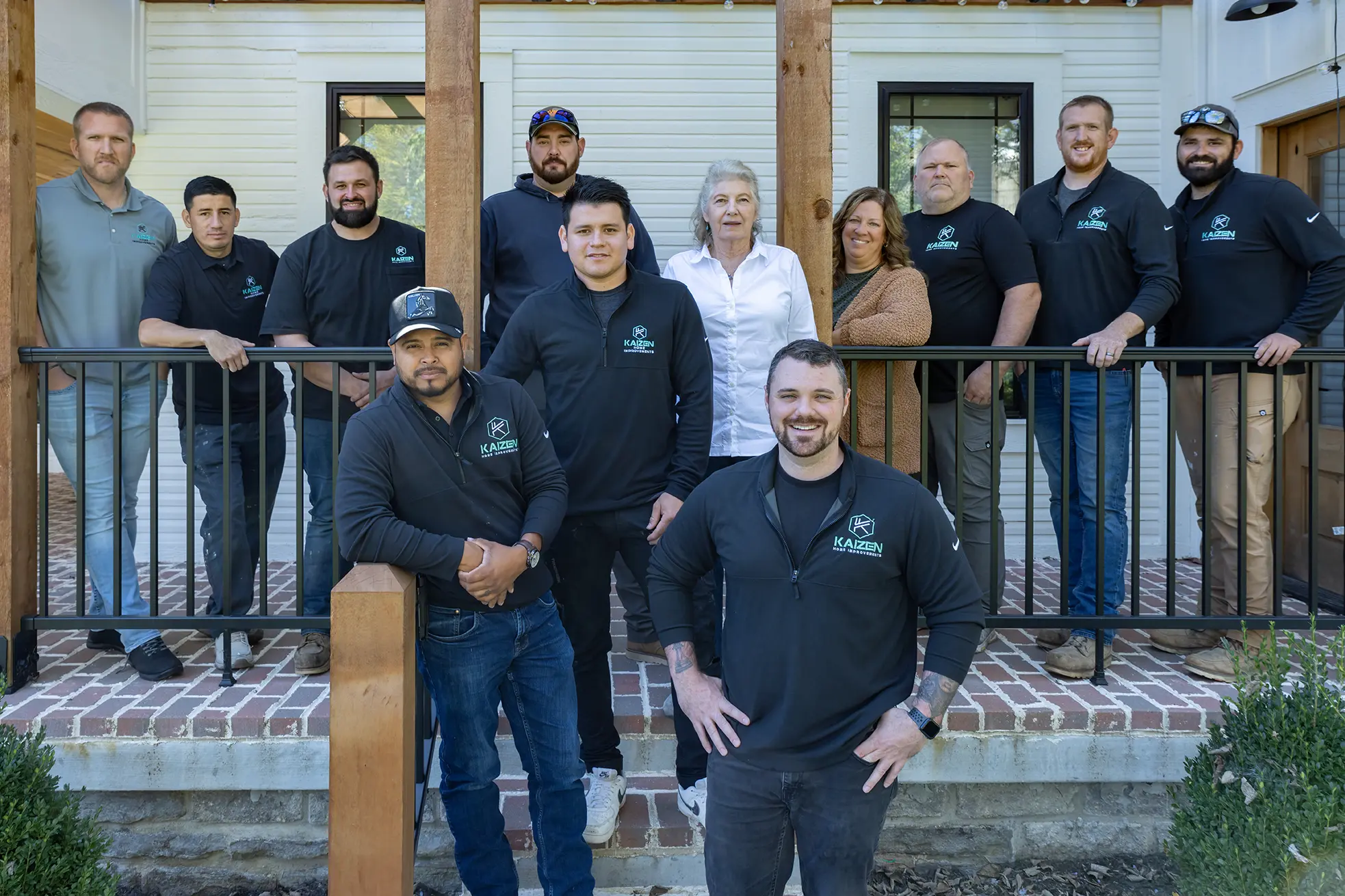 Group of eleven people standing outdoors behind a black metal railing, some smiling, in front of a white building with wooden beams.