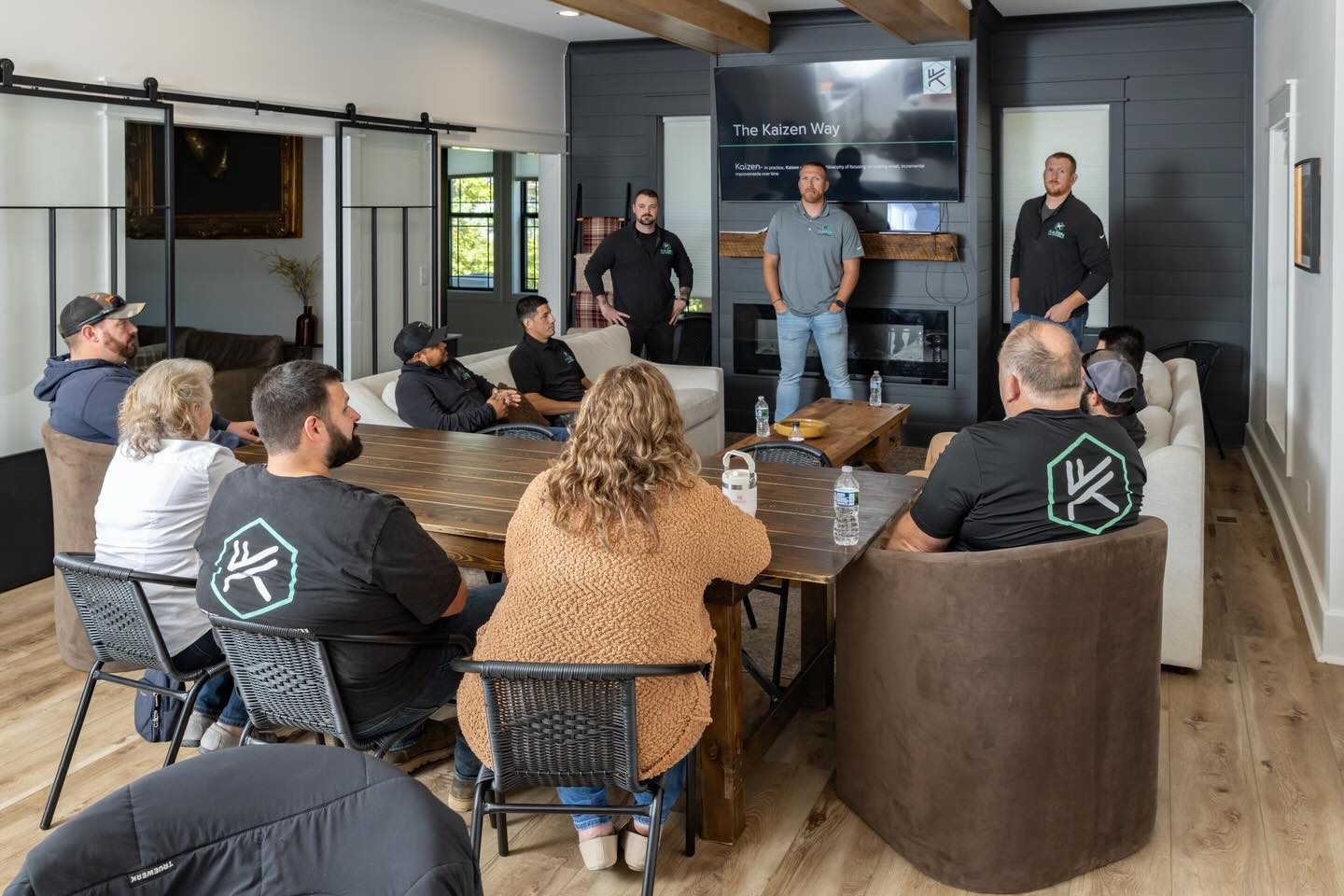 People seated around a table listening to three speakers in a modern room with large windows and a TV screen.