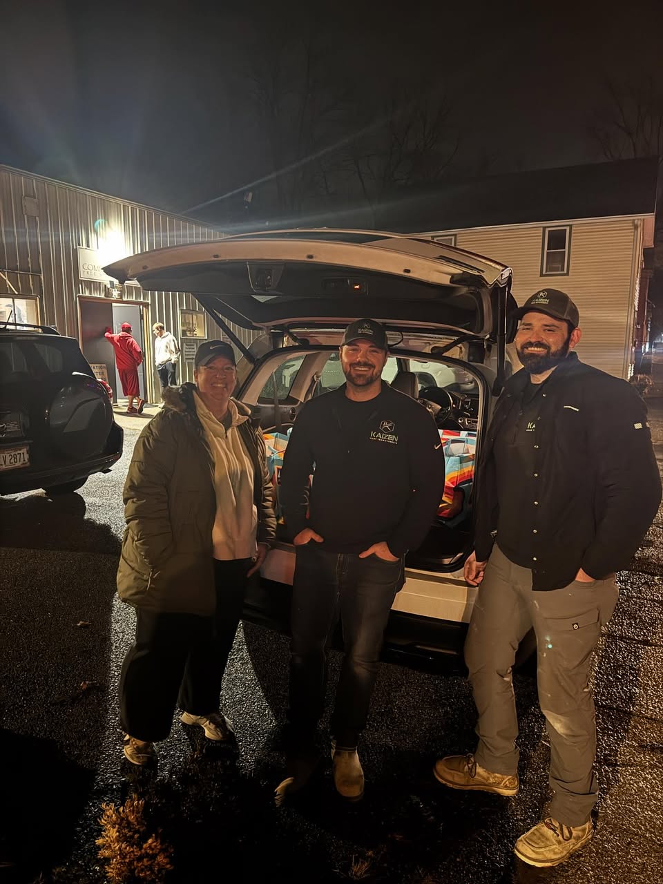 Three people stand outdoors at night in front of an open vehicle trunk, with a building and parked cars behind them.