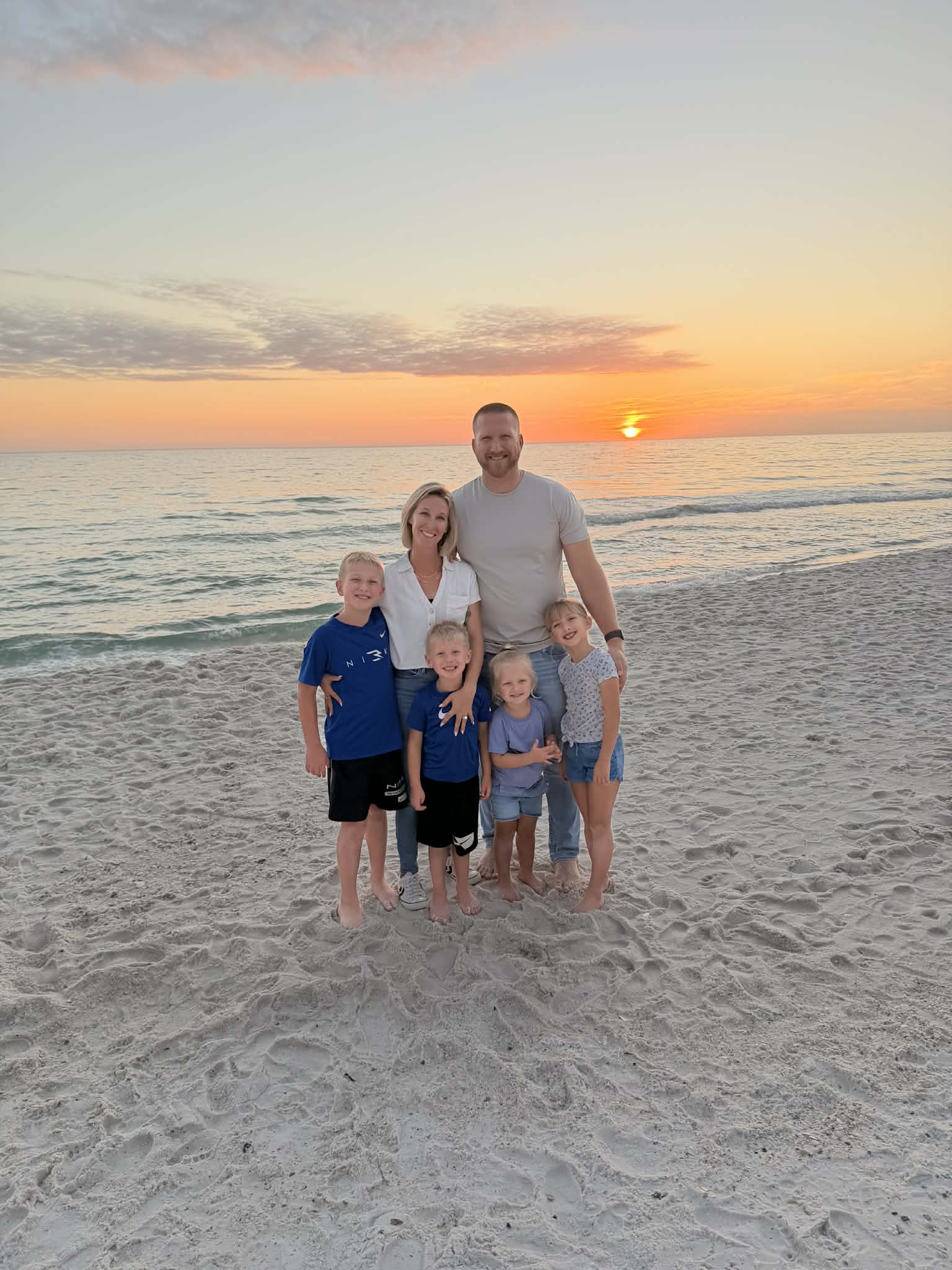 Family of six standing on beach at sunset, smiling, with ocean and sky in background.