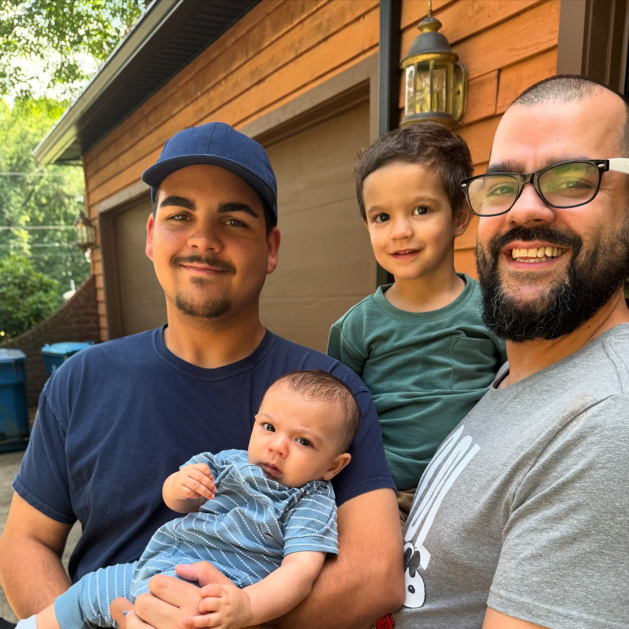 Four people, two adults and two children, smiling outside a house with wooden siding and a lantern.