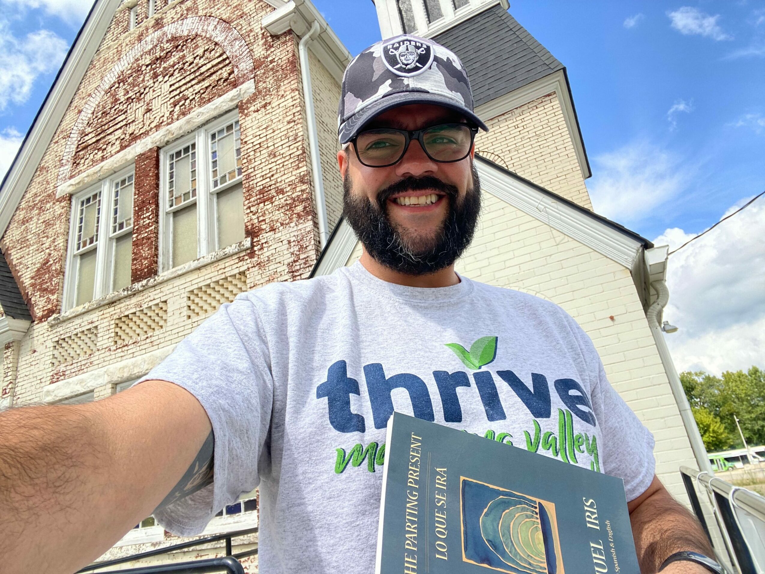 Man wearing sunglasses and a cap, smiling, standing outdoors near buildings and trees.
