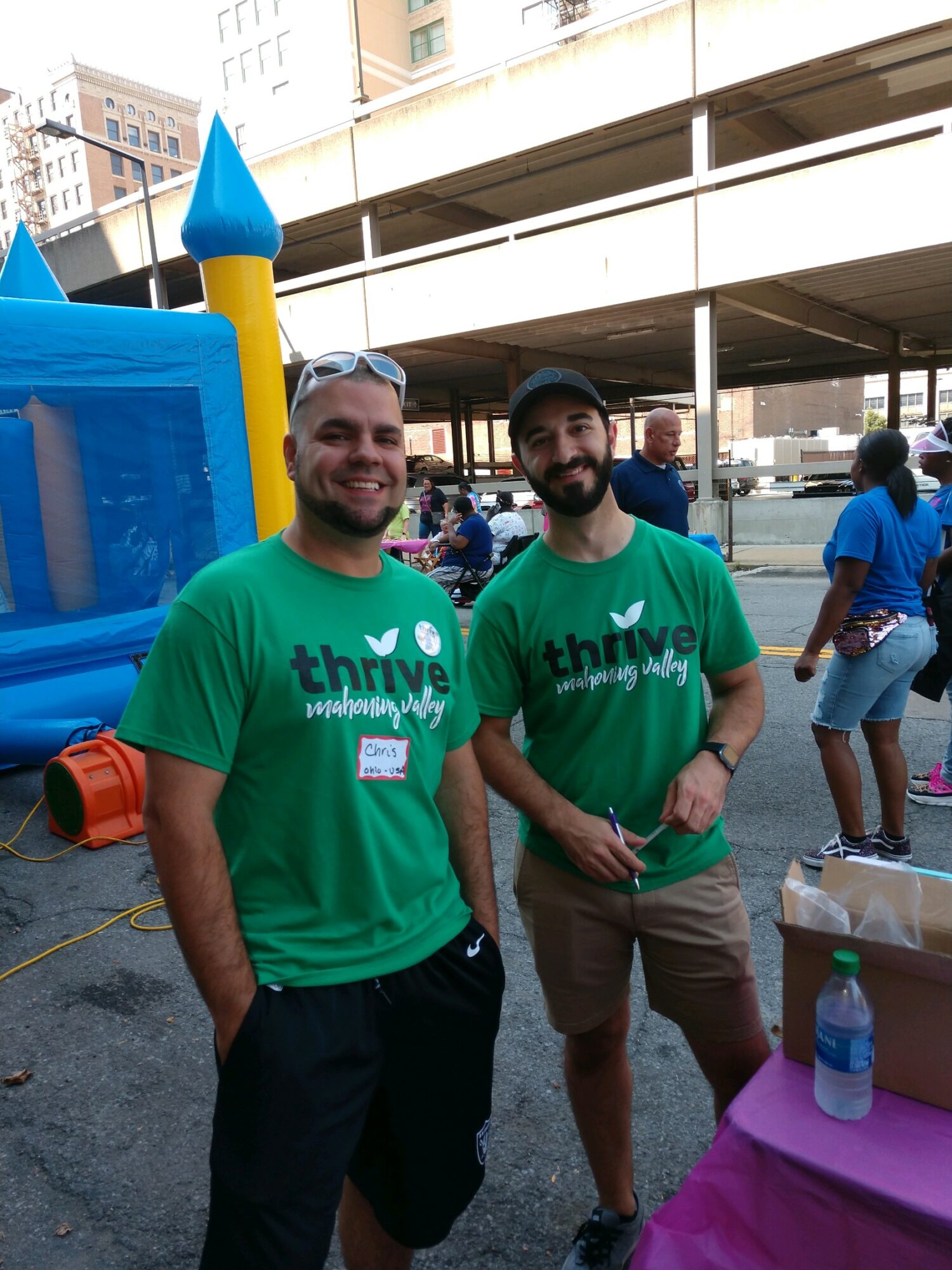 Two smiling men wearing green 'thrive' t-shirts standing outdoors near a table with a water bottle, in an urban setting.