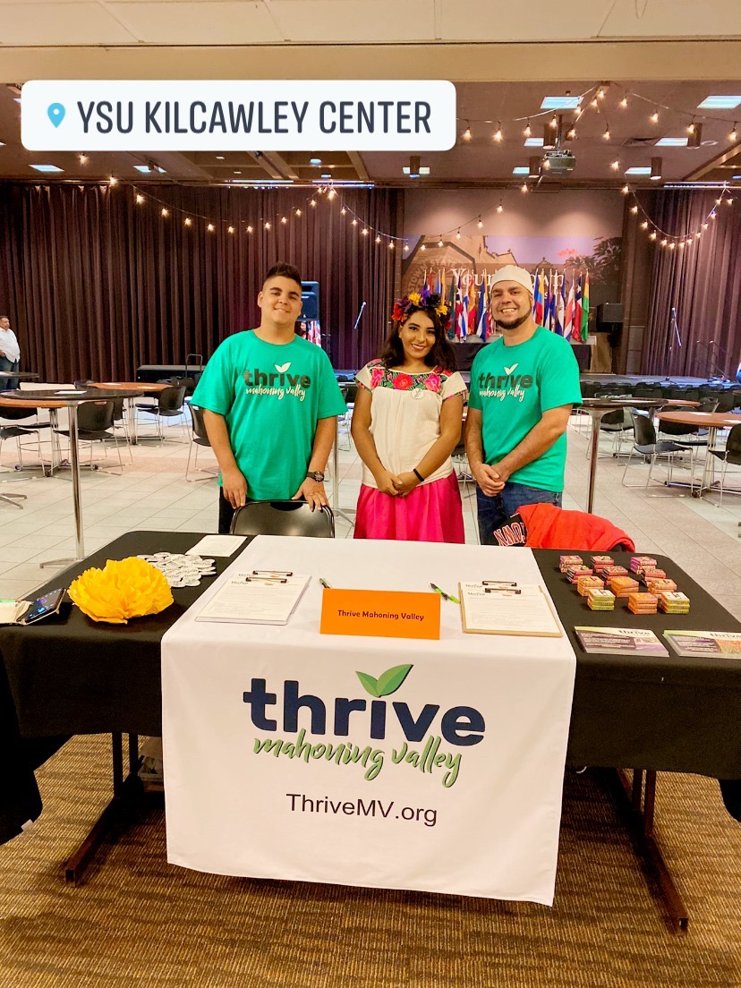 Three people standing behind a table with a Thrive banner, in a large indoor event space with tables and decorations.
