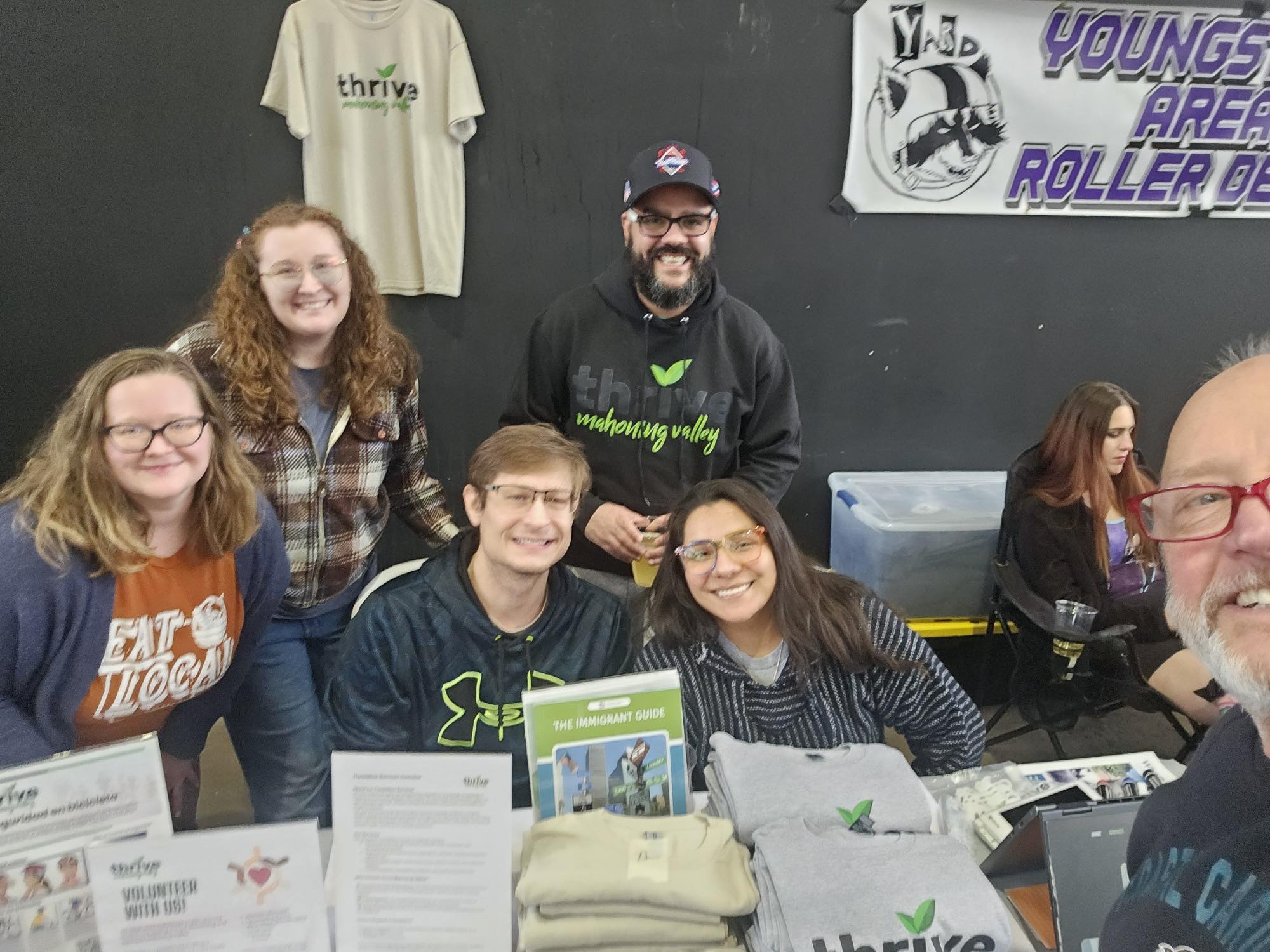 Group of five people sitting and standing at a table, smiling, with a dark wall and posters behind them.