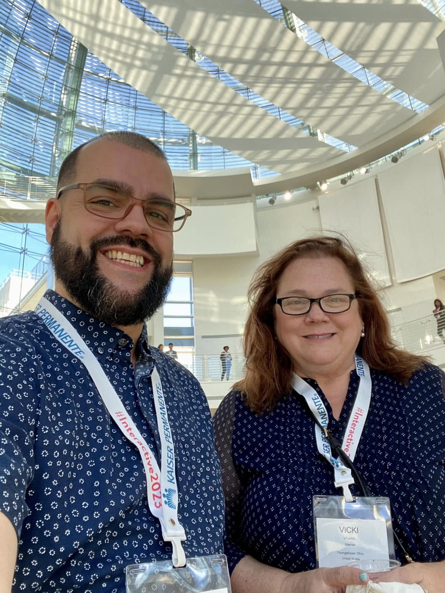 Two people smiling, wearing glasses and conference badges, standing indoors with a modern ceiling structure above.