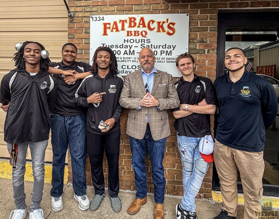 Group of six people standing outside a brick building with a sign for Fat Back's BBQ, smiling and posing for the photo.