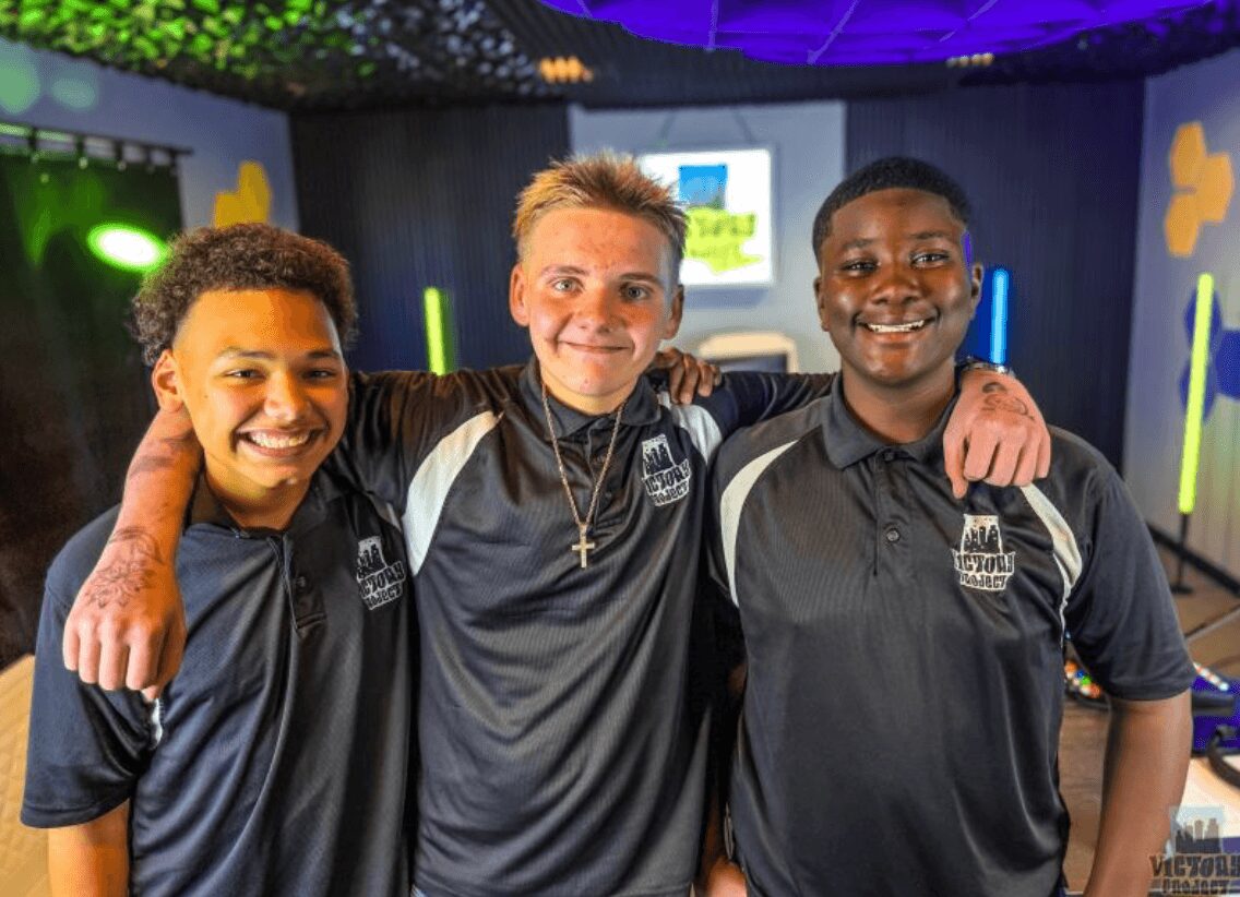 Three smiling young men in matching dark sports shirts with a colorful indoor background.